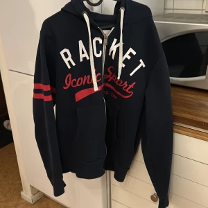 Mörkblå hoodie - Säljer en mörkblå hoodie med texten 'Racket Iconic Sport' i vitt och rött på framsidan. Den har en dragkedja och två röda ränder på ärmen. Perfekt för en stilig lock.