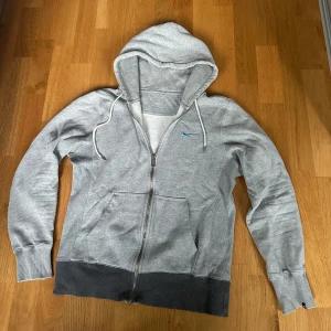 Grå Nike zip hoodie - Säljer en grå hoodie från Nike med dragkedja och blå logga på bröstet. Tröjan har en justerbar huva med snören och är tillverkad i mjukt material för extra komfort. Perfekt för kyliga dagar. S/xs är storleken
