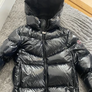Svart pufferjacka från Colmar - Säljer en snygg svart pufferjacka från Colmar. Använd två gånger så nästan helt ny. Jackan har en glansig yta och är utrustad med dragkedja framtill. Perfekt för kyliga dagar med sin vadderade design. Märkeslogga på ärmen ger en extra touch av stil.  Köpt för 4000kr. Passar mellan 150-180 skulle jag säga