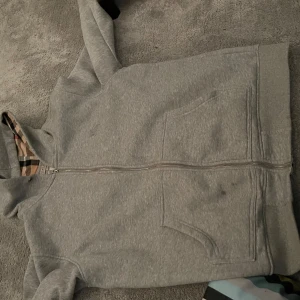 Burberry zip - Säljer en stilren grå huvtröja från Burberry med klassiskt rutigt mönster i huvan. Tröjan har dragkedja framtill och praktiska fickor. Perfekt för en avslappnad look.