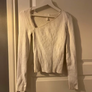 Vit ribbad tröja från H&M - Säljer en stilren vit ribbad tröja från H&M med en unik asymmetrisk halsringning. Tröjan har långa ärmar och är perfekt för lager-på-lager under kyligare dagar. Passar bra till både jeans och kjol för en avslappnad look.