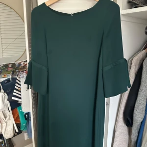 Mörkgrön klänning med volangärmar - Elegant mörkgrön klänning med volangdetaljer på ärmarna. Perfekt för en stilren look. Klänningen har en enkel och klassisk design med rund hals och trekvartsärmar.