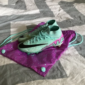 Turkosa fotbollsskor från Nike - Snygga turkosa fotbollsskor från Nike med svart swoosh-logga. Skorna har snörning och en hög ankelkrage för extra stöd. Perfekta för fotbollsplanen med sina dobbar för bra grepp. Kommer med en matchande lila påse med turkosa detaljer.