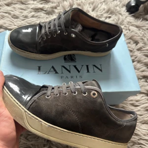 Lanvin skor - Snygga grå sneakers från Lanvin med en kombination av mocka och glansigt material. Skorna har en klassisk snörning och en vit sula med Lanvin-logga undertill. Perfekta för en stilren look. Skor feta nu till våren! Bara att höra av sig vid minsta fundering!