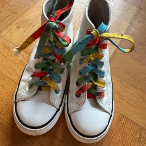 Vita sneakers med färgglada skosnören - Säljer ett par vita sneakers med högt skaft och färgglada skosnören i regnbågens färger. Skorna har en klassisk design med svart rand runt sulan och är perfekta för att ge en färgklick till din outfit. Storlek 33-34
