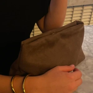 Brun clutch - Snygg brun kuvertväska i mjukt mockamaterial. Perfekt för att bära med sig det viktigaste med stil. Litet hål vid knappen och lite fläckar.Enkel och elegant design som passar till många olika outfits.