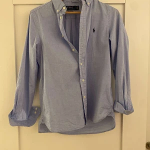 Blå skjorta från Polo Ralph Lauren - Snygg ljusblå skjorta från Polo Ralph Lauren med klassisk krage och knappar framtill. Skjortan har långa ärmar och en broderad logotyp på bröstet. Perfekt för en stilren look!