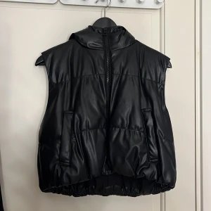 Svart dunväst med dragkedja - Snygg svart dunväst med dragkedja och huva. Perfekt för en cool och avslappnad stil. Västen har en glansig yta och elastisk nederkant för en bekväm passform. Ifrån H&M har storlek S