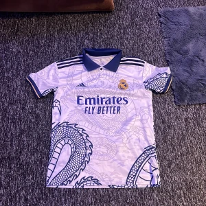 Vit fotbollströja med drakmönster från Adidas - Säljer en vit fotbollströja från Adidas med ett coolt drakmönster. Tröjan har korta ärmar och en mörkblå krage. Den har även klubbens emblem och sponsorlogga på framsidan. Perfekt för fotbollsfans som vill sticka ut!