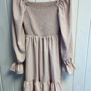 Ljusrosa klänning med smockdetaljer - Ljusrosa klänning med smockdetaljer upptill och långa puffiga ärmar. Klänningen har volanger vid ärmslut och nederkant.💕