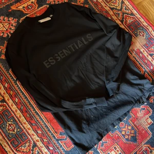 Svart Essentials tröja  - Säljer en stilren svart tröja från Essentials by Fear of God. Tröjan har en klassisk design med långärmade ärmar och en rund halsringning. Perfekt nu när det ska vara lite varmare.