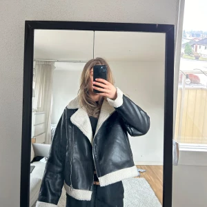Svart skinnjacka med foder - Snygg svart skinnjacka med vit teddyfoder och dragkedja. Jackan har en klassisk design med bred krage och är perfekt för kyligare dagar. Den har en cool och trendig look som passar till många olika stilar.
