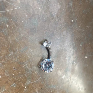 Silverfärgad navelpiercing med glittriga stenar - Snygg navelpiercing i silverfärg med två glittriga stenar. Perfekt för att ge lite extra bling till din stil. Den böjda stången gör den bekväm att bära.