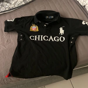 Svart pikétröja från Polo Ralph Lauren - Säljer en svart pikétröja från Polo Ralph Lauren med texten 'CHICAGO' och en stor logotyp på framsidan. Tröjan har en klassisk krage och korta ärmar. På baksidan finns siffran '3' i vitt. Perfekt för en sportig look!