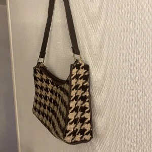 Rutig axelväska i brunt och beige - Snygg axelväska med ett klassiskt rutigt mönster i brunt och beige. Väskan har en praktisk dragkedja och en justerbar axelrem. Perfekt för att ge din outfit en stilfull touch.