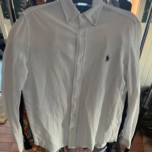 Vit skjorta från Ralph Lauren - Säljer en klassisk vit skjorta från Ralph Lauren med knappar och broderad logga på bröstet. Skjortan har långa ärmar och en stilren design som passar perfekt till olika tillfällen.