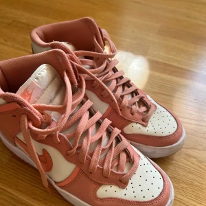Nike sneakers i rosa och vit - Snygga Nike sneakers i en fräsch kombination av rosa och vit. Skorna har en klassisk design med snörning och en högre siluett. Perfekta för att ge din outfit en sportig touch. Nypris 1600kr