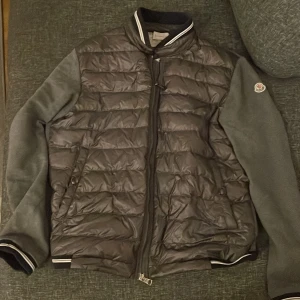 Svart dunjacka från Moncler - Säljer en snygg svart dunjacka från Moncler med grå ärmar och dragkedja framtill. Jackan har en quiltad design och ribbade muddar vid ärmslut och krage. Perfekt för kyligare dagar!