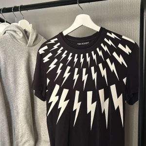 Svart t-shirt med blixtmönster från Neil Barrett - Snygg svart t-shirt från Neil Barrett med ett coolt vitt blixtmönster över hela framsidan. Perfekt för att sticka ut med en unik stil. Kortärmad och i en bekväm passform.