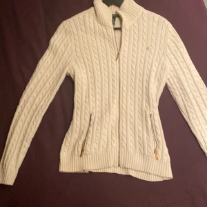 Vit stickad kofta från  Ralph Lauren - Säljer en stilren vit stickad kofta från Ralph Lauren med dragkedja och ribbad krage. Koftan har ett klassiskt flätmönster och praktiska fickor med dragkedja. Perfekt för kyliga dagar! Stl L men passar M