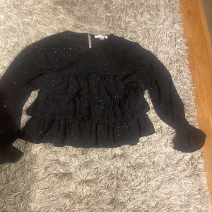 Svart blus från Gina Tricot - Säljer en svart blus från Gina Tricot med glittriga prickar och volanger. Blusen har långa ärmar och en rund halsringning. Perfekt för att lägga till lite glitter i vardagen eller till speciella tillfällen.