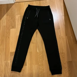 Svart sportbyxa från Lindex - Snygg, svart sportbyxa från barnavdelningen på Lindex i nytt skick, inga defekter! Byxan har mjukt träningstyg och en bekväm passform med snörning i midjan. Byxan har fickor med dragkedja och textdetaljer längs benen. Perfekt för en avslappnad dag eller träning!