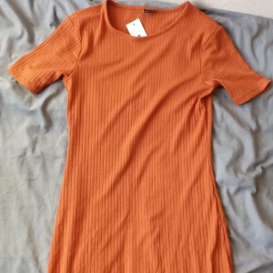 Orange ribbad klänning från Gina Tricot - Säljer en snygg orange ribbad klänning från Gina Tricot. Klänningen har korta ärmar och en liten slits på sidan. Helt nya! Perfekt för en stilren look. Passar bra till både sneakers och klackar. 🧡