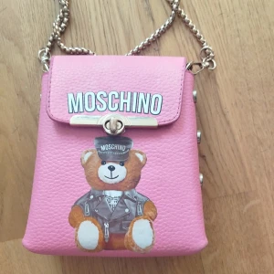 Rosa axelväska från Moschino - Säljer en söt rosa axelväska från Moschino med en kedjerem och ett charmigt nallemotiv på framsidan. Väskan har en tryckknapp och dekorativa nitar på sidan. Perfekt för att ge en lekfull touch till din outfit!