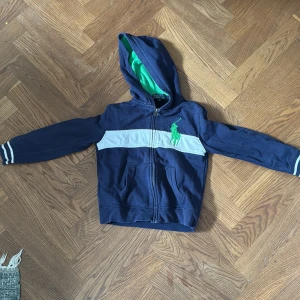 Blå huvtröja från Polo Ralph Lauren - Säljer en blå huvtröja från Polo Ralph Lauren med grönt foder i huvan. Tröjan har en dragkedja framtill. 6 år.