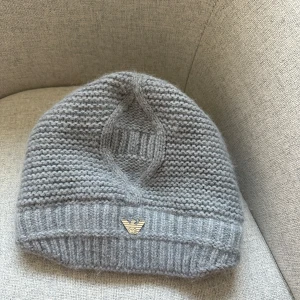 Grå stickad mössa  - Snygg grå stickad mössa med ribbad kant och ett diskret metallmärke framtill. Perfekt för kyliga dagar och ger en stilren look.