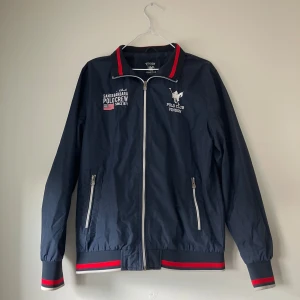 Mörkblå jacka från Vinson Polo Club - Snygg mörkblå jacka från Vinson Polo Club med dragkedja och broderade detaljer. Jackan har långa ärmar och ribbade muddar med röda och vita ränder. Perfekt för en stilren look med en sportig touch.