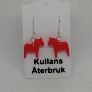 Röda dalahäst örhängen från Kullans Återbruk - Unika örhängen med röda dalahästar från Kullans Återbruk. Perfekta för att lägga till en färgglad och traditionell touch till din stil. Tillverkade med omsorg och kreativitet.