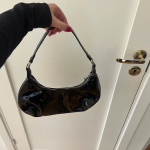 Svart axelväska i syntet - Skit snygg svart väska i glansigt material, perfekt att styla upp en outfit!