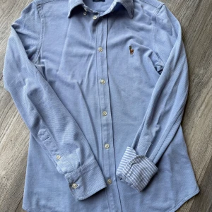 Blå skjorta från Ralph Lauren - Snygg blå skjorta från Ralph Lauren med klassisk krage och broderad logga på bröstet. Skjortan har långa ärmar och knappar framtill. Manschetterna har ett stilrent randigt mönster på insidan för en extra touch.