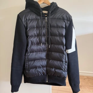 Svart jacka med huva från Moncler - Säljer en snygg svart cardiganfrån Moncler med huva och dragkedja. Jackan har en quiltad framsida och långa ärmar med ett stort vitt M på ärmen. Perfekt för kyligare dagar och även lite varma dagar.Säljer den då jag inte använder den.Det är storlek m men passar även om du har storlek s 