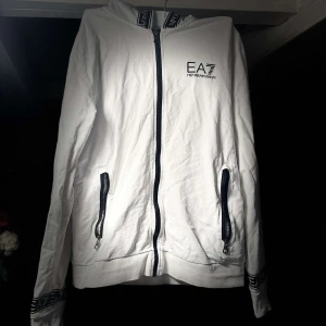 Vit huvtröja från EA7 - Säljer en stilren vit huvtröja från EA7 med dragkedja och svarta detaljer. Tröjan har långa ärmar och ribbade muddar. Perfekt för en avslappnad look.
