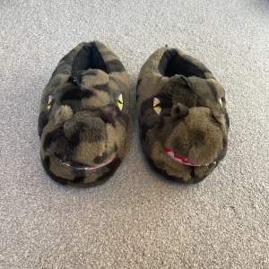 Varma tofflor som liknar dinosaurier - Mysiga tofflor i form av dinosaurier med kamouflagemönster. De har broderade ögon och en rolig design med tänder som ger dem ett lekfullt utseende. Perfekta för att hålla fötterna varma och bekväma hemma. Stl 37-39. Aldrig använda