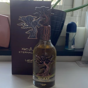 Eternal Oud Parfym från Lattafa - Elegant parfymflaska med ett unikt trädmotiv i bronsfärg på både flaska och lock. Flaskan är genomskinlig och innehåller en gyllene vätska. Förpackningen är mörkbrun med guldfärgad text. Perfekt för den som söker en distinkt och lyxig doftupplevelse.95ml kvar