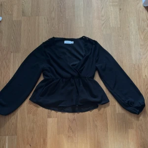 Svart omlottblus från Vila - Säljer en elegant svart omlottblus från Vila. Blusen har långa ärmar och en smickrande omlottdesign som ger en fin siluett. Perfekt för att klä upp eller ner beroende på tillfälle. 🖤