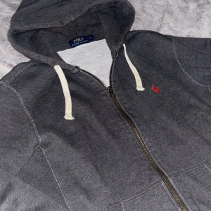 Grå hoodie från Polo Ralph Lauren - Säljer en grå hoodie från Polo Ralph Lauren med vit snörning i huvan och röd broderad logga på bröstet. Tröjan har dragkedja framtill, fickor och ribbade muddar. Perfekt för chill dagar och streetwear-stil. Pris är ej hugget i sten