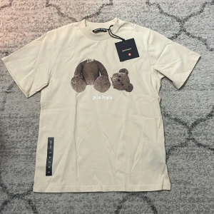 Beige Palm Angels t-shirt med nalletryck - Snygg beige t-shirt från Palm Angels i bomull med ett stort tryck av en brun nalle på framsidan. Klassisk rund hals och korta ärmar. Palm Angels-loggan är tryckt under motivet. Perfekt för dig som gillar streetwear med en twist.