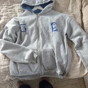 Grå hoodie med blå broderi och dragkedja - Grå hoodie med blå broderade bokstäver på bröstet, hel dragkedja framtill och två fickor. Insidan av huvan är blå, vilket ger en snygg kontrast. Tröjan har långa ärmar och ribbade muddar vid ärmslut och nederkant. Perfekt för chill dagar. Obs. Lappen är borta så ser inte storleken men skulle gissa på en M