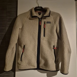 Beige teddyjacka från Patagonia XL - Säljer en beige teddyjacka från Patagonia i storlek XL även storlek 14 i barn storlek. Jackan har lång ärm, hel dragkedja framtill och tre fickor med snygga kontrastdetaljer i rött. Mjuk och fluffig fleece som håller dig varm och chill under kalla dagar. Priset ej hugget I sten.