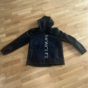 Svart Moncler Windbreaker - Svart vindjacka från Moncler med huva och stora vita logotypbokstäver framtill. Jackan har dragkedjor på fickorna och är helsvart med Moncler-märke på ärmen. Materialet är polyester och jackan har en clean, sportig look med lång ärm och normal passform.