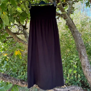 Svart långklänning från H&M Mama - Svart långklänning från H&M Mama med smock upptill och rak, luftig kjol. Klänningen är axelbandslös och har en enkel, stilren design som passar perfekt för varma dagar. Materialet är mjukt och känns skönt mot huden. Snygg att styla med sneakers eller sandaler. Storlek M, men passar en L. Fint skick, men fortfarande mycket kvar att ge. 