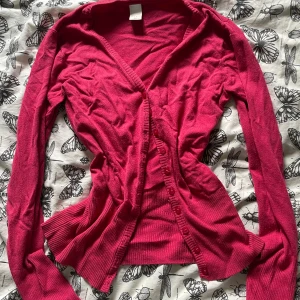 Rosa kofta med knappar - Säljer en snygg rosa kofta med v-ringad hals och långa ärmar. Koftan har ribbade muddar och knappar framtill, perfekt att slänga över en t-shirt eller linne. Mjuk och skön, passar dig som gillar färg och vill ha en enkel men stilren look.