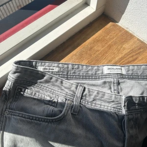 Grå jeans från Jack & Jones - Snygga grå jeans från Jack & Jones med klassisk femficksdesign och ljus tvätt. Modellen har raka ben och normal passform, perfekt för dig som gillar en enkel och stilren look. Tillverkade i slitstarkt bomullsmaterial med detaljerade sömmar och läderpatch bak.