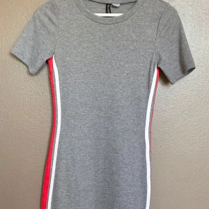 H&M Divided grå t-shirtklänning sportig randig strl 38/M - Sportig och bekväm t-shirtklänning från H&M Divided.   Grå färg med röda och vita ränder längs sidorna, figurnära passform och kort ärm.    Storlek: 38/M Skick: Mycket fint skick.   Nypris ca 199 kr.    Perfekt till vardag – snygg med sneakers eller jeansjacka.