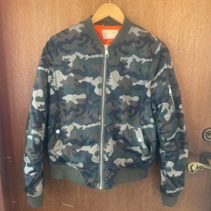 Camouflage bomberjacka med dragkedja - Snygg bomberjacka med klassiskt camouflage-mönster i gröna, grå och svarta toner. Jackan har ribbade muddar, dragkedja framtill och fickor med tryckknappar. Orange foder på insidan ger en cool kontrast. Perfekt för dig som vill sticka ut med streetstyle.