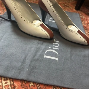 Dior pumps i vitt och brunt skinn - Snygga pumps från Dior i vitt och brunt skinn med spetsig tå och hög klack. Skorna har dekorativa perforerade detaljer längs kanterna och en elegant brun panel framtill. Perfekt för dig som gillar klassisk stil med en twist.